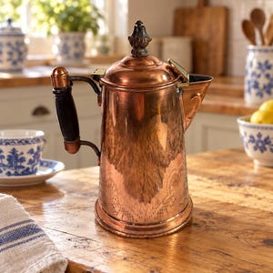 May include: A polished copper coffee pot featuring a cylindrical body with a subtle embossed lace-like pattern, a dark wooden handle with metallic accents, a hinged lid with a decorative finial, and a wide pouring spout on the side.