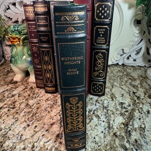 May include: A collection of antique books with decorative gold lettering and accents. The spines are various colors, including dark green, burgundy, and black. The book titled "Wuthering Heights" is prominently displayed.