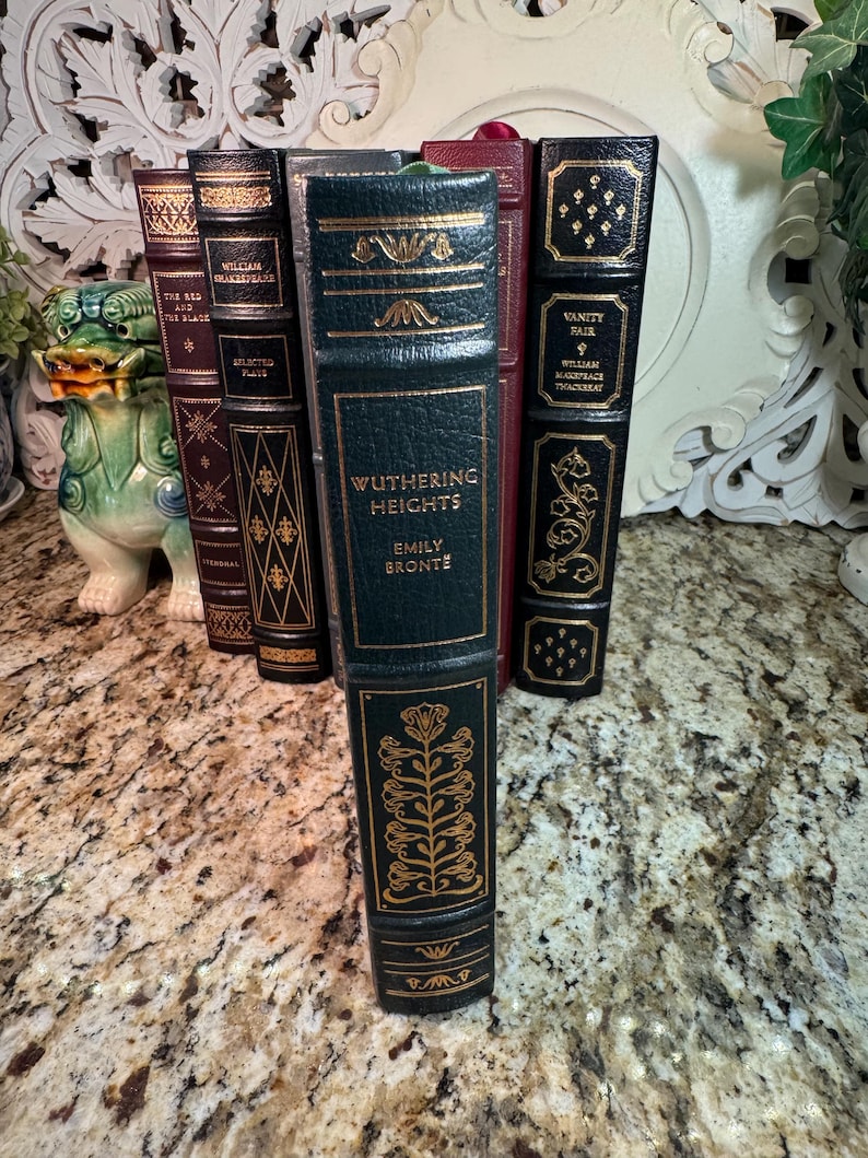 May include: A collection of antique books with decorative gold lettering and accents. The spines are various colors, including dark green, burgundy, and black. The book titled "Wuthering Heights" is prominently displayed.