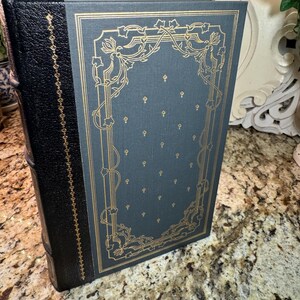 May include: A hardcover book with a black leather spine and a teal cover. The cover features gold floral designs and small gold accents. The book's pages are bound with a red ribbon.