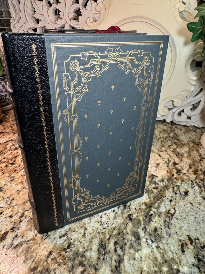 May include: A hardcover book with a black leather spine and a teal cover. The cover features gold floral designs and small gold accents. The book's pages are bound with a red ribbon.