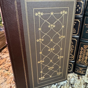 May include: A hardcover book with a brown leather spine and a gray cover featuring a gold floral design. The book's edges are trimmed in gold. Other books are visible in the background.