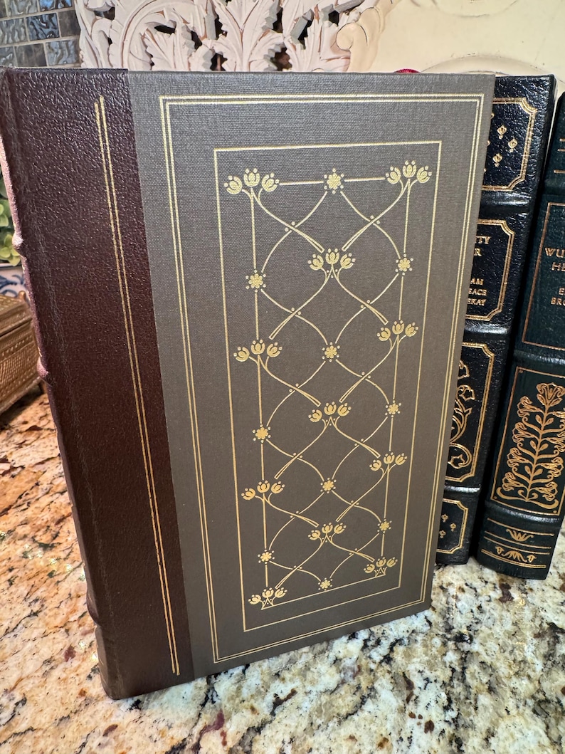 May include: A hardcover book with a brown leather spine and a gray cover featuring a gold floral design. The book's edges are trimmed in gold. Other books are visible in the background.
