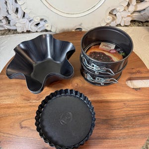 May include: Three black metal baking pans on a wooden surface. The pans are different sizes and shapes. The largest pan is a round springform pan with a removable bottom. The middle pan is a fluted tart pan. The smallest pan is a fluted tart pan with a removable bottom.