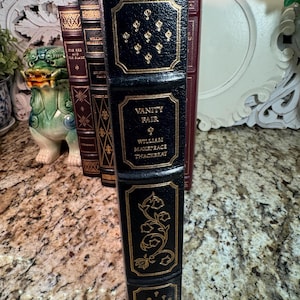 May include: A black leather-bound book titled "Vanity Fair" by William Makepeace Thackeray, with gold embossed details. The spine features decorative gold accents and text. Other books are visible in the background.