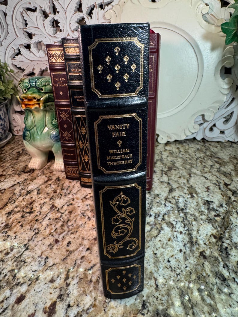 May include: A black leather-bound book titled "Vanity Fair" by William Makepeace Thackeray, with gold embossed details. The spine features decorative gold accents and text. Other books are visible in the background.