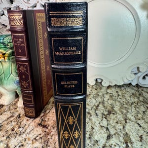 May include: A collection of antique books with decorative gold accents. The spine of the William Shakespeare book is black with gold lettering and ornate designs. The other books have burgundy and brown covers.