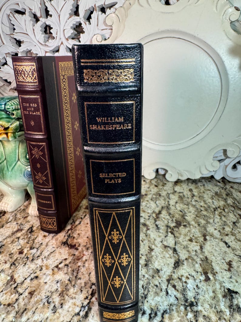 May include: A collection of antique books with decorative gold accents. The spine of the William Shakespeare book is black with gold lettering and ornate designs. The other books have burgundy and brown covers.
