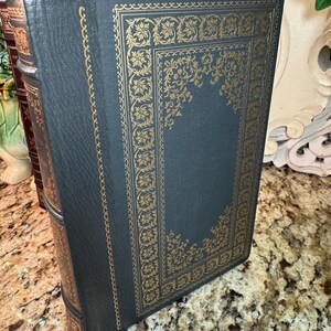 May include: A dark teal book with gold floral detailing. The cover features a gold rectangular border with ornate floral designs. The spine and edges are also detailed in gold. The book is standing upright, showcasing its decorative cover.