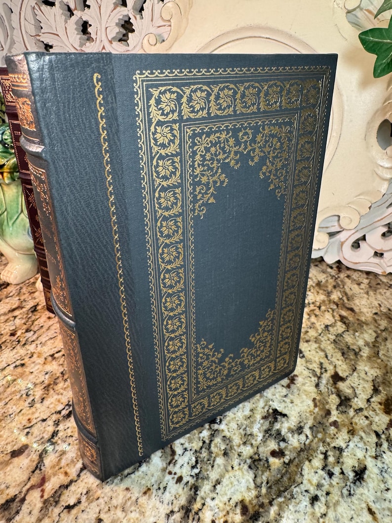 May include: A dark teal book with gold floral detailing. The cover features a gold rectangular border with ornate floral designs. The spine and edges are also detailed in gold. The book is standing upright, showcasing its decorative cover.