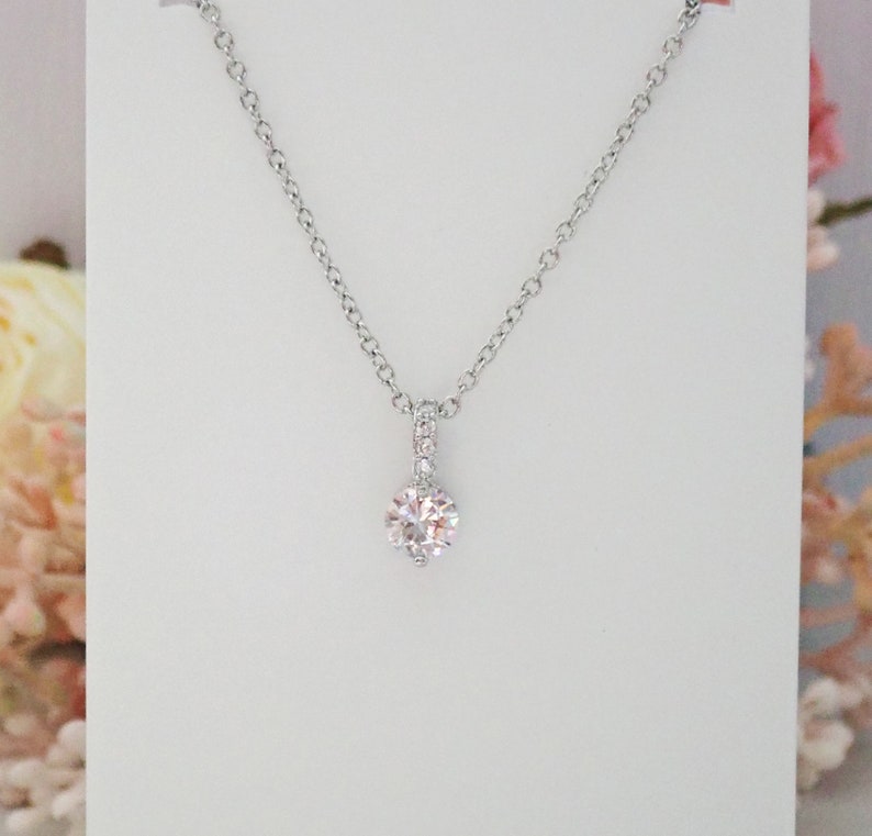 May include: A silver-toned necklace featuring a delicate chain and a single round, clear, faceted stone pendant. The pendant is suspended from a small, vertical bail that is embellished with tiny, sparkling clear stones for added detail.