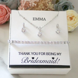 May include: A jewelry set featuring a silver-toned necklace with a round clear stone pendant, matching drop earrings, and a tennis bracelet with clear stones. The set is displayed on a white card with the text EMMA and THANK YOU FOR BEING MY Bridesmaid!