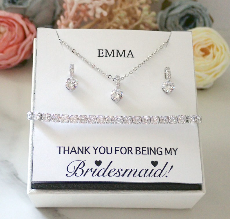 May include: A jewelry set featuring a silver-toned necklace with a round clear stone pendant, matching drop earrings, and a tennis bracelet with clear stones. The set is displayed on a white card with the text EMMA and THANK YOU FOR BEING MY Bridesmaid!