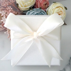 May include: A square white gift box with a textured surface, wrapped in a smooth, wide white satin ribbon tied into a large, centered bow. The box is positioned in front of a cluster of artificial cream, pink, and blue fabric flowers.