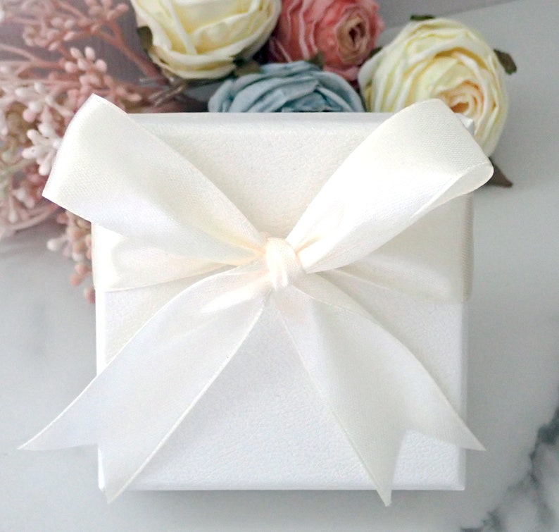 May include: A square white gift box with a textured surface, wrapped in a smooth, wide white satin ribbon tied into a large, centered bow. The box is positioned in front of a cluster of artificial cream, pink, and blue fabric flowers.