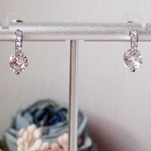 May include: A pair of silver-toned drop earrings featuring a circular clear gemstone suspended from a small hoop. The hoop is embellished with a row of tiny, sparkling white stones. The earrings are displayed on a silver-toned metal stand.