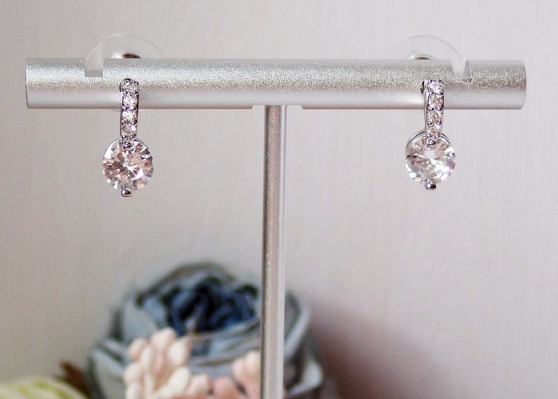 May include: A pair of silver-toned drop earrings featuring a circular clear gemstone suspended from a small hoop. The hoop is embellished with a row of tiny, sparkling white stones. The earrings are displayed on a silver-toned metal stand.