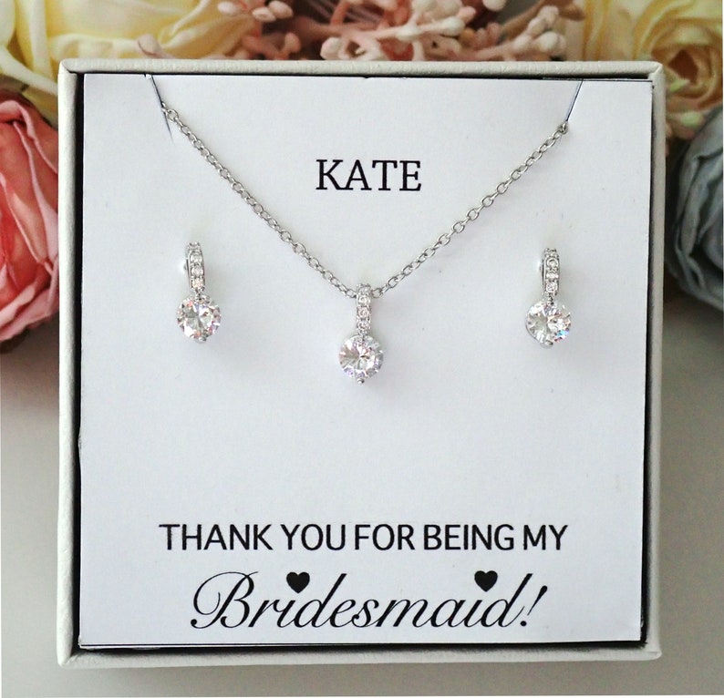 May include: A silver-toned necklace and earring set featuring clear round stones on a white display card. The card displays the text KATE and THANK YOU FOR BEING MY Bridesmaid! with two small black heart icons between the words.