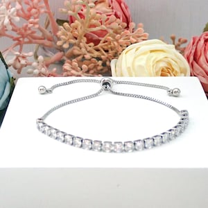 May include: A silver-toned adjustable slider bracelet featuring a row of clear, square-cut stones. The bracelet has a delicate chain with a sliding bead mechanism for size adjustment, finished with two small silver-toned metal beads at the ends.