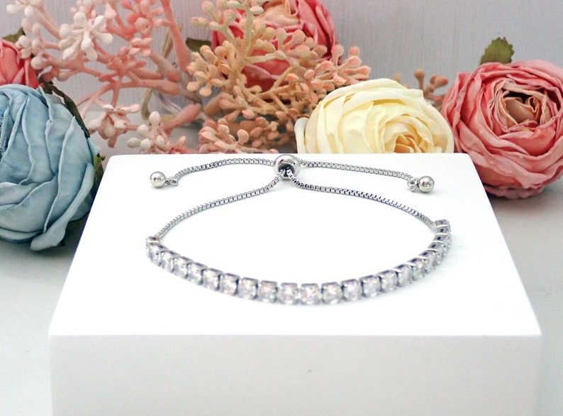 May include: A silver-toned adjustable slider bracelet featuring a row of clear, square-cut stones. The bracelet has a delicate chain with a sliding bead mechanism for size adjustment, finished with two small silver-toned metal beads at the ends.