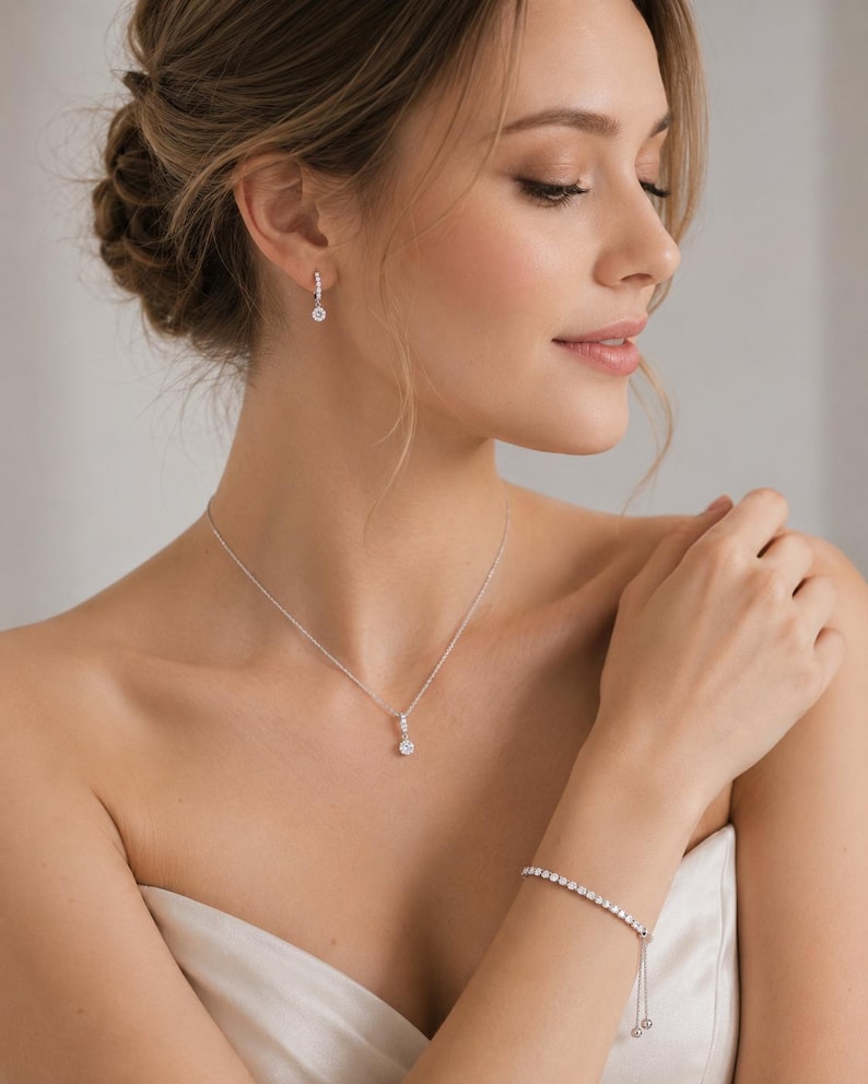 May include: A silver-toned jewelry set consisting of a delicate chain necklace with a single round clear stone pendant, a matching tennis-style bracelet with an adjustable sliding clasp, and small hoop earrings featuring a dangling round clear stone.