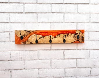 May include: A wooden wall-mounted coat rack with four black hooks. The rack features a natural wood grain design with a vibrant orange resin river. The rack is mounted on a white brick wall.