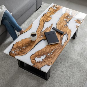 May include: A rectangular coffee table with a white epoxy resin surface and natural wood grain patterns. The table has black legs and a glossy finish. A coffee cup, remote, and notebook rest on the table.