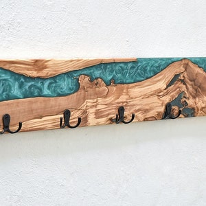 Può includere: Un attaccapanni da parete in legno con un design a fiume in resina epossidica turchese. Il legno ha una venatura naturale ed è accentuato da cinque ganci in metallo nero. L'attaccapanni è montato su una parete bianca.