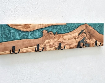 May include: A wooden wall-mounted coat rack with a teal epoxy resin river design. The wood is light brown with dark grain patterns. Five black metal hooks are attached to the bottom edge.
