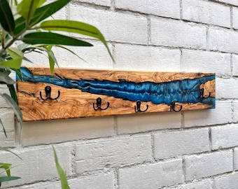 May include: A wooden wall-mounted coat rack with a river of blue resin. The wood has a natural grain pattern. Four black metal hooks are attached to the bottom. The rack is mounted on a white brick wall.