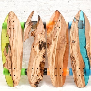 Könnte beinhalten: Vier individuelle Skateboards mit Holzdecks und farbigen Harzakzenten. Jedes Board hat ein einzigartiges Holzmaserungsmuster und ist auf Achsen mit grünen, orangefarbenen und blauen Rollen montiert. Die Boards werden vor einer weißen Backsteinmauer präsentiert.