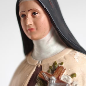 Può includere: Statuina religiosa vintage di una suora, con velo nero e abito color crema. La statuina tiene in mano una croce con una figura argentata di Gesù ed è adornata con rose bianche e foglie verdi. La statua ha un volto dipinto con un'espressione serena.