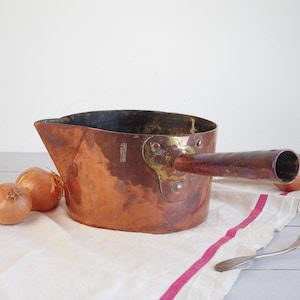 May include: A vintage copper saucepan with a wooden handle, resting on a white cloth with a pink stripe. Two onions sit beside the pan. A silver fork is also present.