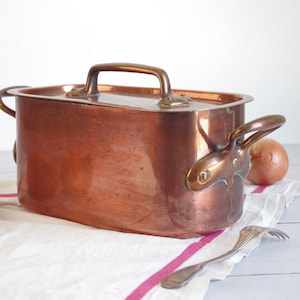 Puede incluir: Una olla rectangular de cobre con tapa y dos asas laterales, más un asa superior. La olla está sobre un paño blanco con una raya rosa, junto a dos cebollas y un tenedor.