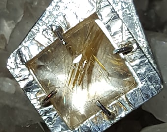 May include: A silver pendant featuring a clear quartz crystal with gold inclusions. The pendant is shaped like a triangle with a square opening in the centre. The pendant is displayed on a bed of white crystals.