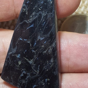 May include: A dark blue and black gemstone cabochon with a swirling pattern. The gemstone is cut in a triangular shape with rounded edges.