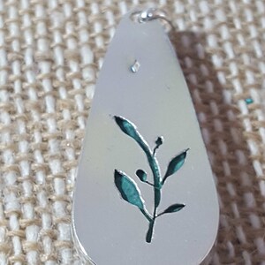 May include: A silver pendant in the shape of a teardrop with a cutout design of a green leafy branch.