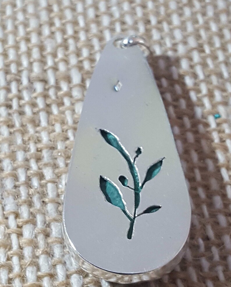 May include: A silver pendant in the shape of a teardrop with a cutout design of a green leafy branch.