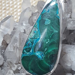 May include: A teardrop-shaped pendant featuring a green and blue gemstone set in silver. The gemstone has a swirling pattern and is surrounded by a silver bezel.