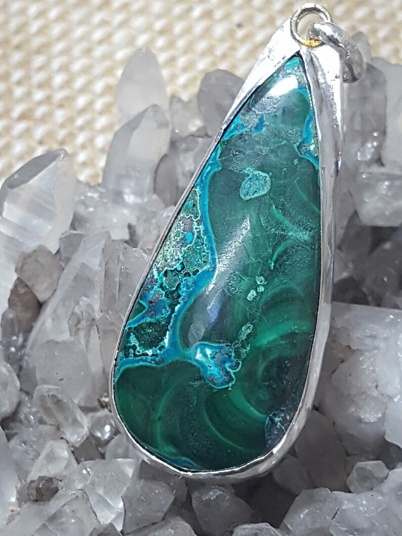 May include: A teardrop-shaped pendant featuring a green and blue gemstone set in silver. The gemstone has a swirling pattern and is surrounded by a silver bezel.
