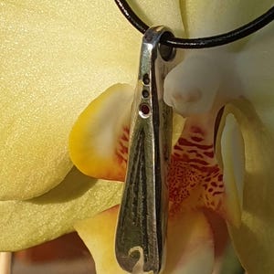 Peut inclure: Un pendentif en argent de forme allongée et étroite avec une surface texturée. Le pendentif est orné de trois petites pierres précieuses rouges disposées en rangée. Le pendentif est suspendu à un cordon en cuir noir.