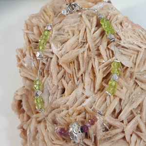 Peut inclure: Un bracelet en argent avec des perles vertes et roses. Le bracelet a un fermoir en argent et un charme en argent au centre.