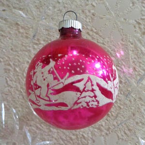 May include: A vintage pink Christmas ornament with a white winter scene featuring skiers and a snowy landscape. The ornament has a silver cap and a loop for hanging. A classic holiday decoration.