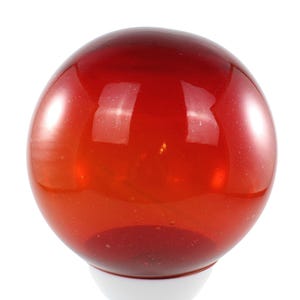 Könnte beinhalten: Ein runder, transparenter, roter Glasgegenstand mit weißem Sockel. Die Glaskugel hat eine glänzende Oberfläche und scheint ein dekorativer Gegenstand zu sein. Die rote Farbe ist reich und lebendig, und das Objekt ist vor einem weißen Hintergrund platziert.