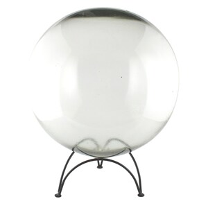 May include: A clear glass sphere, possibly a decorative gazing ball, sits on a dark metal tripod stand. The sphere is transparent, allowing a view through it. The stand has three curved legs that meet at a central point.