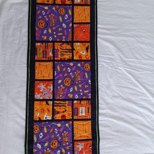 May include: A black and green Halloween table runner with a patchwork design featuring various Halloween-themed fabrics. The fabrics include orange, purple, and black colours with images of pumpkins, candy, ghosts, and other Halloween motifs.