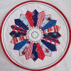 May include: A round, white quilted table topper with a red and blue starburst design. The design features various fabrics with red, white, and blue patterns, including gingham, stripes, and a London bus print.