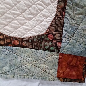 Peut inclure: Gros plan sur un coin de courtepointe avec un motif de patchwork. La courtepointe est faite de diff&eacute;rents tissus dans des tons de brun, bleu et vert. Le tissu a un motif batik avec de petits points et des volutes. La courtepointe est matelass&eacute;e avec un motif en losange.