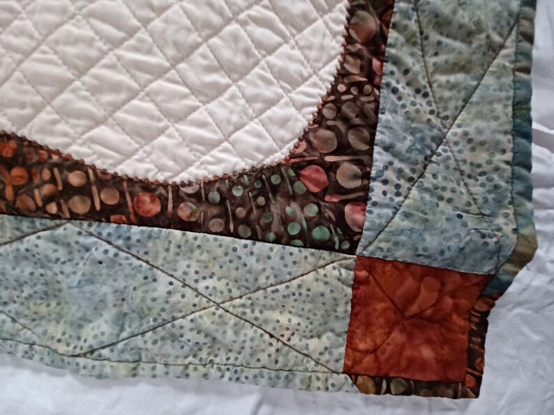 Peut inclure: Gros plan sur un coin de courtepointe avec un motif de patchwork. La courtepointe est faite de diff&eacute;rents tissus dans des tons de brun, bleu et vert. Le tissu a un motif batik avec de petits points et des volutes. La courtepointe est matelass&eacute;e avec un motif en losange.