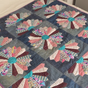Op de afbeelding: Een quilt gemaakt van denim en kleurrijke stofresten. De quilt heeft een herhalend patroon van grote, veelkleurige windmolens.